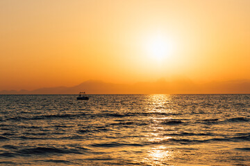 Obraz premium Majestic golden sunset over the ocean with a small silhouetted boat resting on the horizon. Bright sun, shimmering reflection across the rolling waves, peaceful and romantic summer atmosphere.