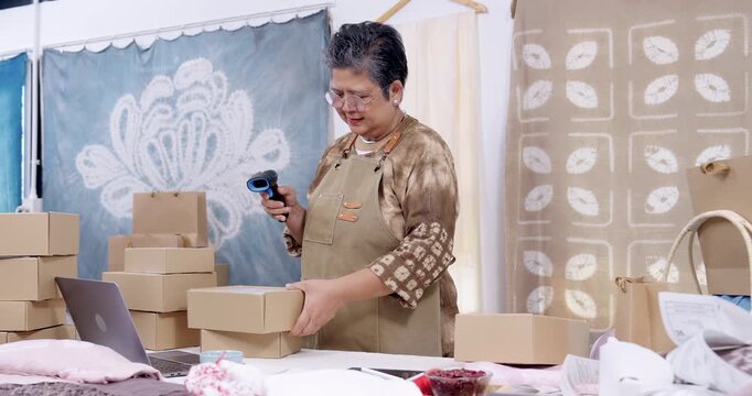 Retired businesswoman lifts carton, scans barcode sticker, checks invoice slip, watches laptop order status, rearranges parcel stack, prepares courier pickup from home studio workspace.