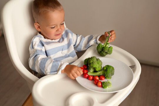 A young child is sitting in a high chair. The child is holding broccoli and looking at cherry tomatoes on a plate. The setting is a simple home kitchen