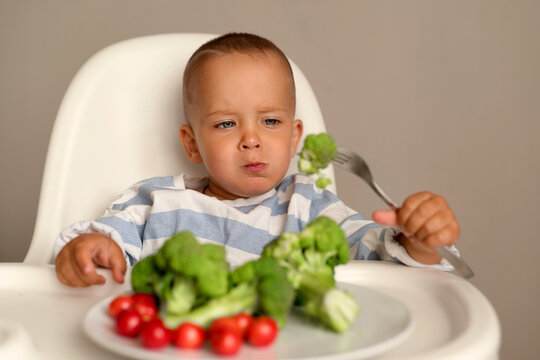 A young child sits in a high chair with broccoli and tomatoes on the plate. The child holds a fork and looks at the food with a thoughtful expression
