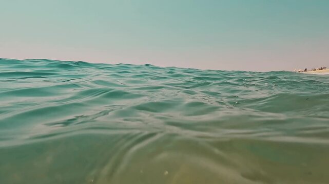 V&iacute;deo grabado a ras de agua en mar abierto, con la superficie del mar en primer plano, horizonte bajo y una mujer nadando a lo lejos. La escena tiene una est&eacute;tica minimalista y serena, con tonos suave