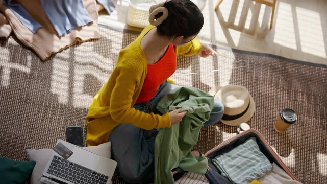 High angle view of a young Asian woman packing clothes into an open suitcase. Female traveler preparing luggage, choosing outfits, and getting ready for a holiday vacation trip. Travel preparation.