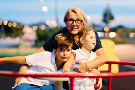 Grandmother With Two Grandchildren Riding a Playground Merry-Go-Round at Dusk, Family Moment
