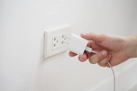 close up hand plugging adapter charge the battery with outlet socket plug and hold mobile phone on wall ,electric safety concept