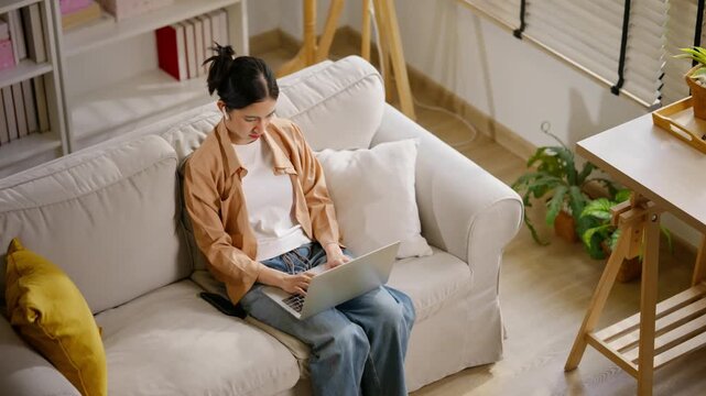 Young Asian woman working on laptop computer while sitting on a cozy white sofa in her living room. Female freelancer or remote worker typing on a laptop. working from home, digital lifestyle concept.