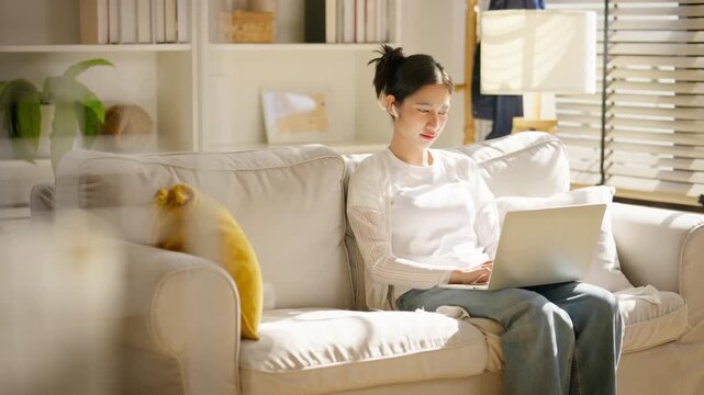 Young Asian woman working on laptop computer while sitting on a cozy white sofa in her living room. Female freelancer or remote worker typing on a laptop. working from home, digital lifestyle concept.