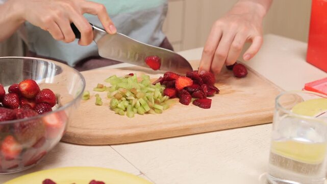 Woman's hands chopping strawberries and rhubarb vegetable stems on a wooden cutting board, preparing ingredients for baking, jam, or a healthy dessert in a home kitchen
