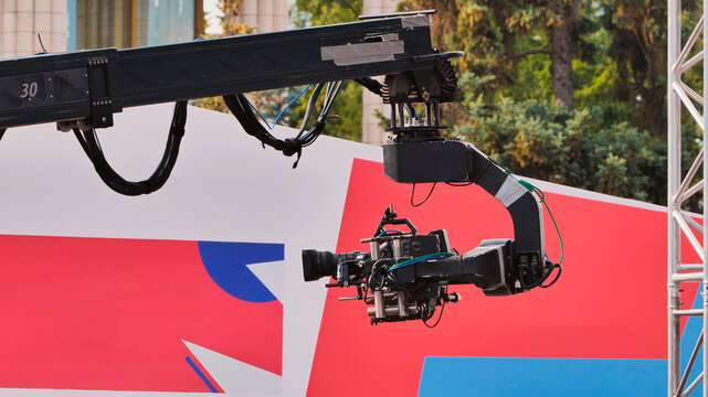 Large television camera on a jib crane at an outdoor broadcast event with a colorful background