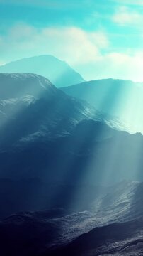 Light beams shine through a mountain range during early morning. The scene shows layers of mountains with a hazy atmosphere and soft light illuminating the landscape.