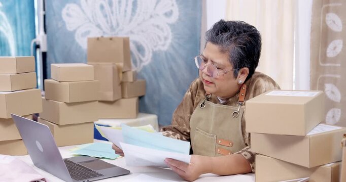 Senior businesswoman reviews paperwork stack, checks laptop order management, compares invoice slip with receipt note, plans parcel packing steps, prepares courier pickup inside home studio.