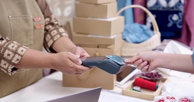 Asian senior businesswoman in apron holds POS terminal while customer taps credit card, confirms cashless payment, checks invoice receipt, stacks parcel box for e-commerce shipping in home studio.