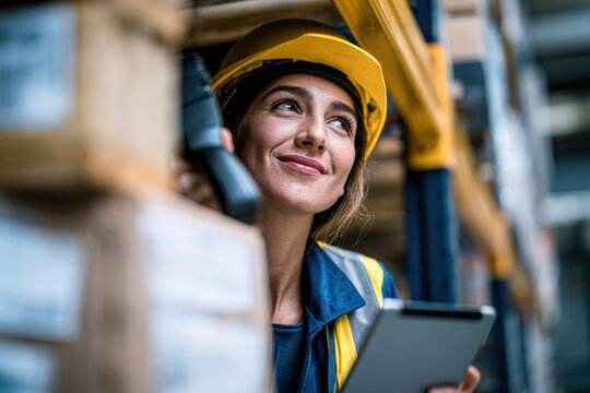 A female worker wearing a yellow hard hat and safety vest holds a digital tablet while inspecting inventory in a warehouse filled with stacked boxes.