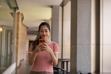 Fototapeta premium Young Asian Woman Smiling While Using Smartphone Outdoors in Modern Building Corridor