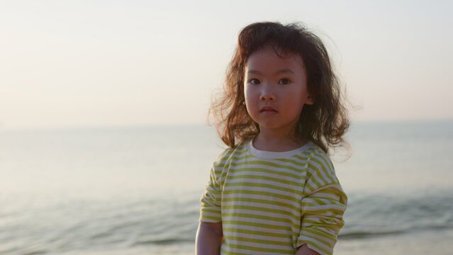 A young Asian girl with a curious expression stands on the beach at sunset, looking into the camera and embodying a sense of quiet childhood contemplation.