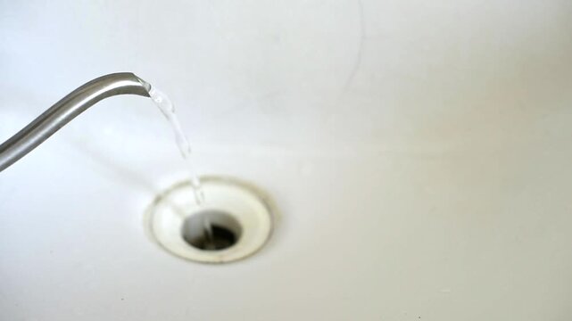 A close-up shot shows hot water being poured from a stainless steel kettle spout directly into a white ceramic sink drain. The steady stream flows into the hole to clear a clog.