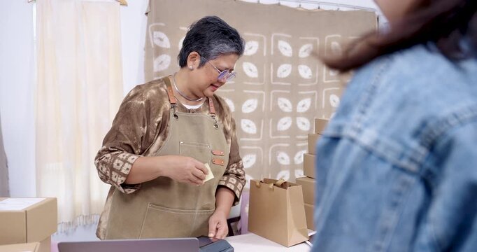 Asian senior entrepreneur delivers parcel bag to customer after payment, confirms order on laptop, updates paperwork processing, organizes boxes for small business shipping in home studio workspace.