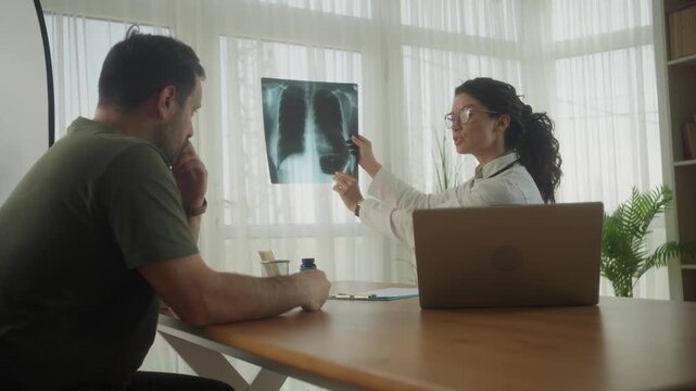 Confident female doctor showing chest x-ray image to male patient and explaining diagnosis during checkup in clinic
