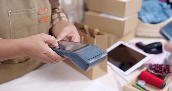 Slow motion closeup, retired asian woman seller presents EDC machine, customer taps phone for contactless payment, stacked parcels wait, small business order shipping continues inside home studio.