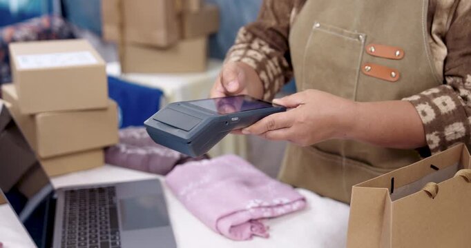 Elderly asian artisan online seller tilts POS terminal toward customer phone, payment approval icon appears, laptop business management runs behind, shibori textile parcels queued for delivery.