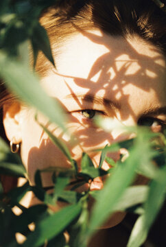 A woman gazes through green leaves dappled in shadow and light