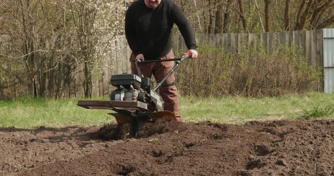 Farmer using a rototiller to plow a vegetable patch in the backyard, getting the land ready for spring planting. A man operating a walk-behind tractor to cultivate the soil
