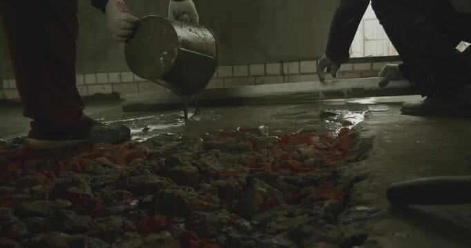 Construction workers pouring liquid cement from a bucket onto a rough surface and leveling the wet screed during a house renovation, laying the foundation for a new floor on the building site