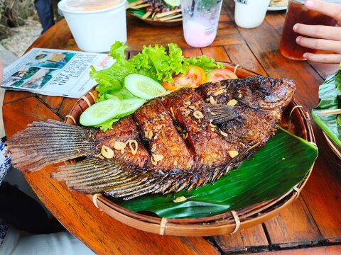 Traditional dish of crispy fried gourami fish served on banana leaf in a bamboo woven basket complete with fresh lettuce, cucumber, and tomato.