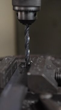Close-up of a high-speed steel drill bit drilling a hole into a metal plate held by a vise, creating metal shavings and swarf in an industrial workshop