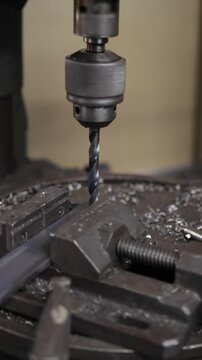 Close-up of a drill press drilling a hole in a metal bar held by a vise, with metal shavings flying and a worker lubricating the drill bit for precision work