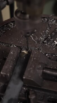 Close-up of a drill press machine boring a hole into a metal bar held in a vise, creating shavings and smoke in an industrial manufacturing workshop or factory setting