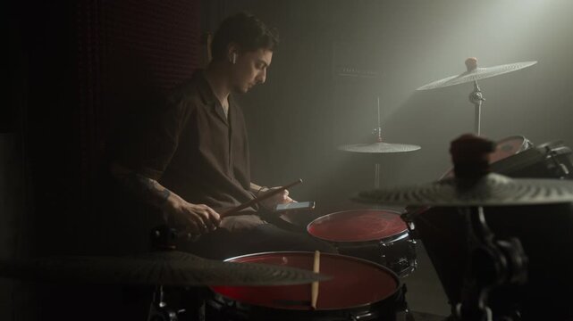Caucasian young man sitting at drum kit, using smartphone and wearing wireless earbuds. Footage for promoting music learning apps, online drum lessons, or audio equipment