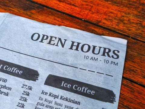 Information on coffee shop operational hours reading open hours 10 AM to 10 PM on a menu sheet over a brown wooden table.