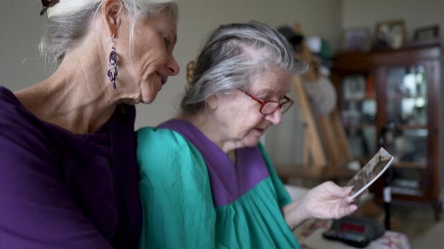A mature daughter visits her elderly mother at a senior care facility. They share stories and look at old photos together during the visit.