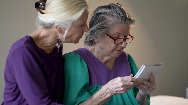 Mature daughter visits elderly mother at senior care facility. They share stories and look at photos together. Moments of connection occur.