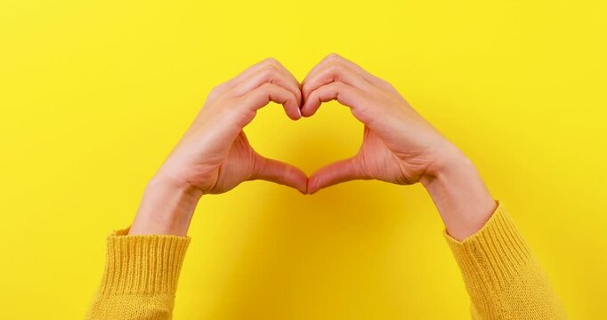 Close-up video of hands forming a heart shape, separating, and coming together again on a bright yellow background. Concept of love, connection, relationships, unity, care, and affection.