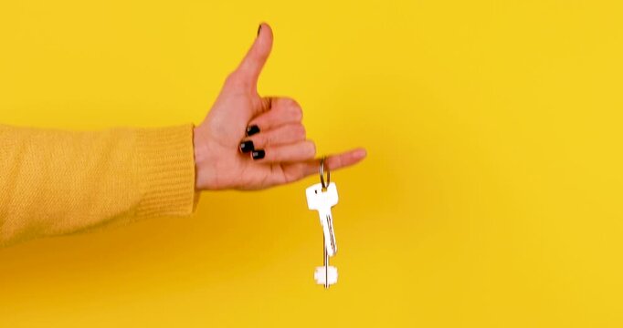 Close-up video of house or apartment keys hanging from a finger while a hand gently sways on a bright yellow background. Real estate concept of home ownership, property purchase, rental, and moving.