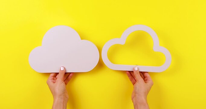 Close-up video of hands holding two cloud symbols on a bright yellow background. Concept of cloud storage, cloud computing, data synchronization, backup, file sharing, and digital technology.