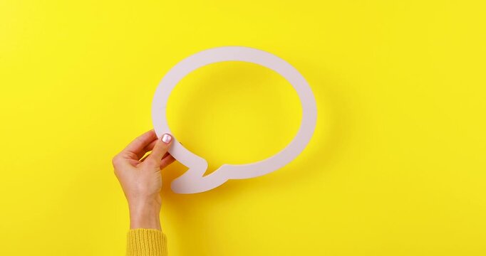 Close-up video of a hand holding a shaking 3D speech bubble on a bright yellow background. Concept of messaging, notifications, chat, communication, social media, and instant messages.