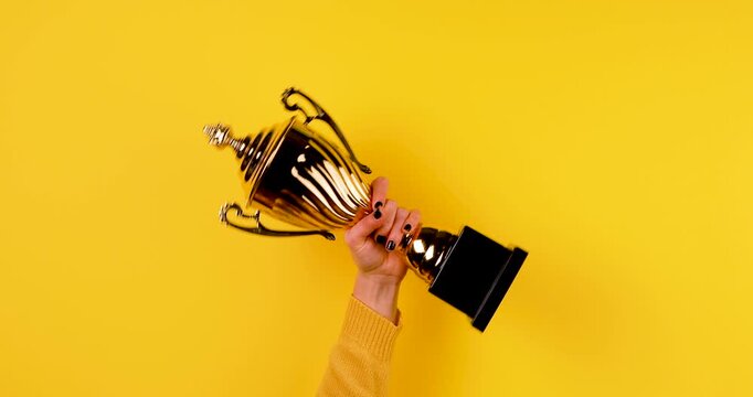 Close-up video of a hand raising and shaking a golden winner trophy on a bright yellow background. Concept of victory, success, achievement, championship, celebration, and first place.