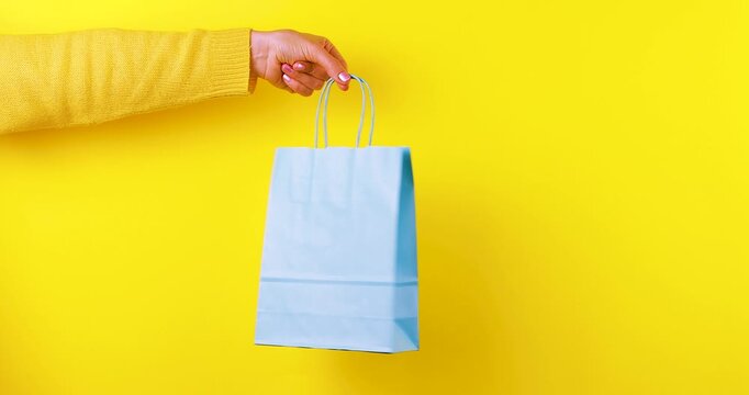 Close-up video of a hand holding and rotating a blue paper shopping bag on a bright yellow background. Retail and shopping concept for stores, gifts, sales, branding, packaging, and e-commerce.
