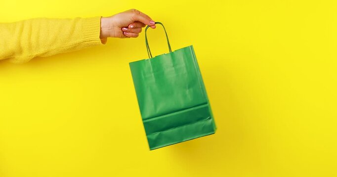 Close-up video of a hand holding and rotating a green paper shopping bag on a bright yellow background. Eco-friendly shopping concept symbolizing sustainability, reusable packaging, and green