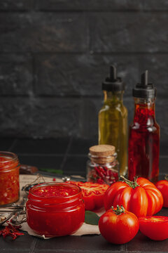 Homemade tomato sauces, ketchup and adjika in glass jars with heirloom tomatoes on dark background
