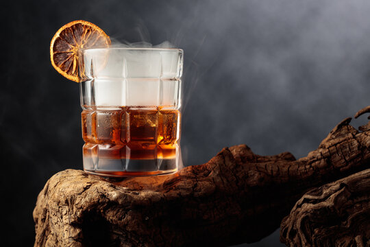 Smoked old-fashioned cocktail with ice and a dried orange slice served on weathered driftwood.