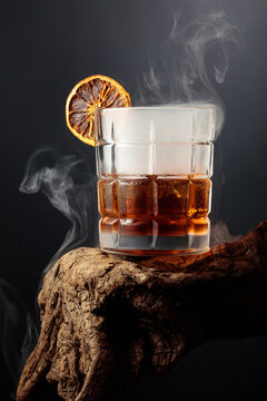 Smoked old-fashioned cocktail with ice and a dried orange slice served on weathered driftwood.