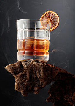 Smoked old-fashioned cocktail with ice and a dried orange slice served on weathered driftwood.