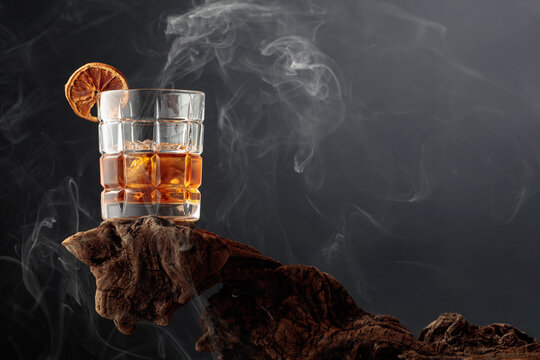 Smoked old-fashioned cocktail with ice and a dried orange slice served on weathered driftwood.