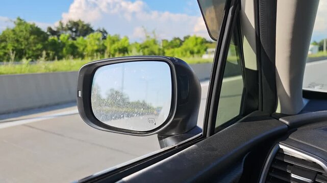 Passenger point of view showing the side mirror of a car moving along a highway. Sunny day with trees and blue sky visible in the background