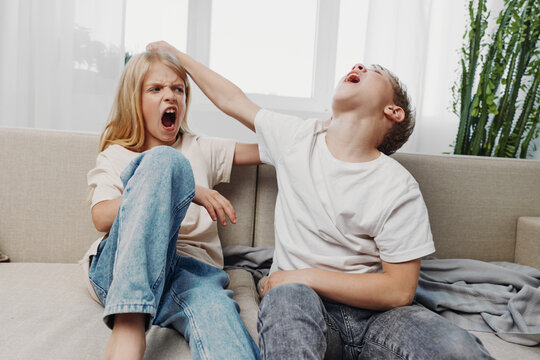Two children in casual clothes sit on a couch indoors, one boy playfully pulling a girl&rsquo;s hair while she reacts with a frustrated expression. Concept of sibling rivalry and childhood conflict.