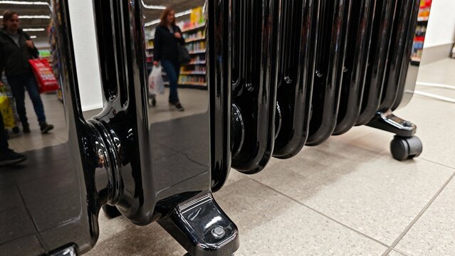A sleek black radiator sits quietly on the floor, reflecting light and surroundings. People browse aisles filled with colorful goods. The atmosphere buzzes with energy in a warm, inviting space.