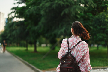 Naklejka premium Young person outdoors, walking away in a park, striped shirt, backpack, breeze in hair, sunlit path, vibrant greenery, casual mood, candid travel moment, relaxed posture, fresh air, subtle motion